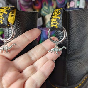 Könnte beinhalten: Ein Paar schwarze Lederstiefel mit gelb-schwarzen Fersenschlaufen, die den Schriftzug AirWair tragen. Jeder Stiefel ist mit einem silberfarbenen Metallring an der Schlaufe versehen, an dem ein kleiner silberfarbener Dinosaurier-Anhänger hängt.