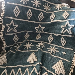 K&ouml;nnte beinhalten: Eine tealblaue geh&auml;kelte Wolldecke mit wei&szlig;em Pompon-Saum. Die Decke zeigt ein sich wiederholendes Muster aus Schneeflocken, Diamanten und Weihnachtsb&auml;umen.