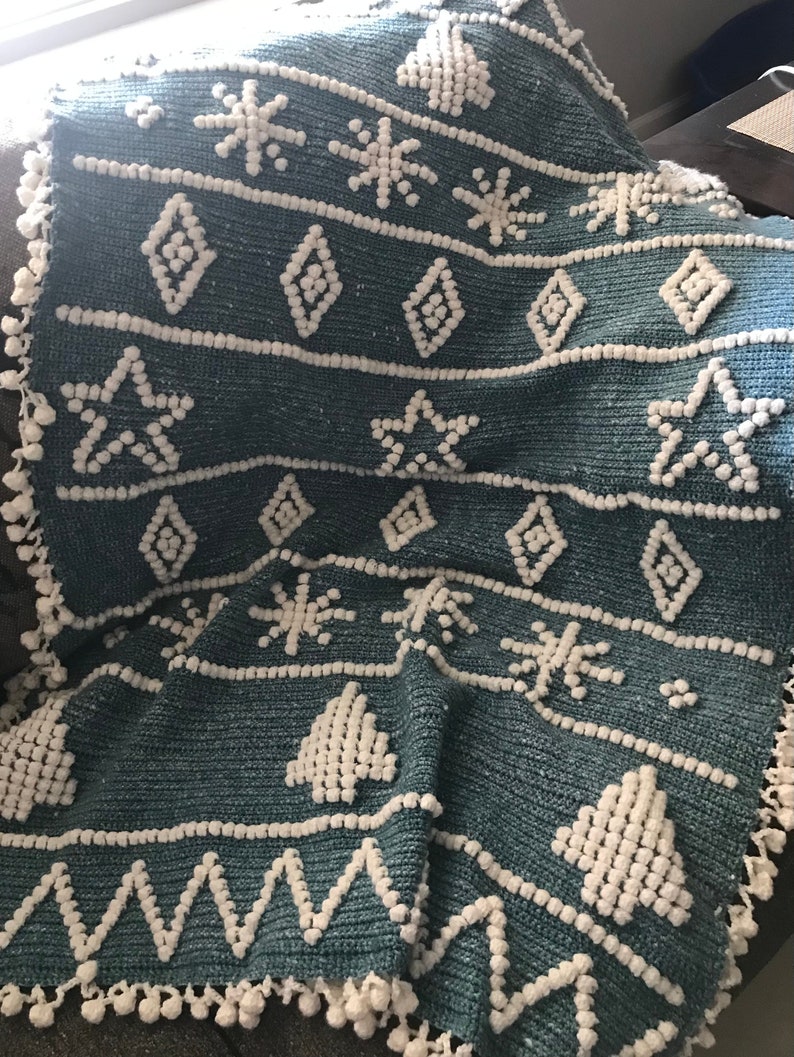 K&ouml;nnte beinhalten: Eine tealblaue geh&auml;kelte Wolldecke mit wei&szlig;em Pompon-Saum. Die Decke zeigt ein sich wiederholendes Muster aus Schneeflocken, Diamanten und Weihnachtsb&auml;umen.