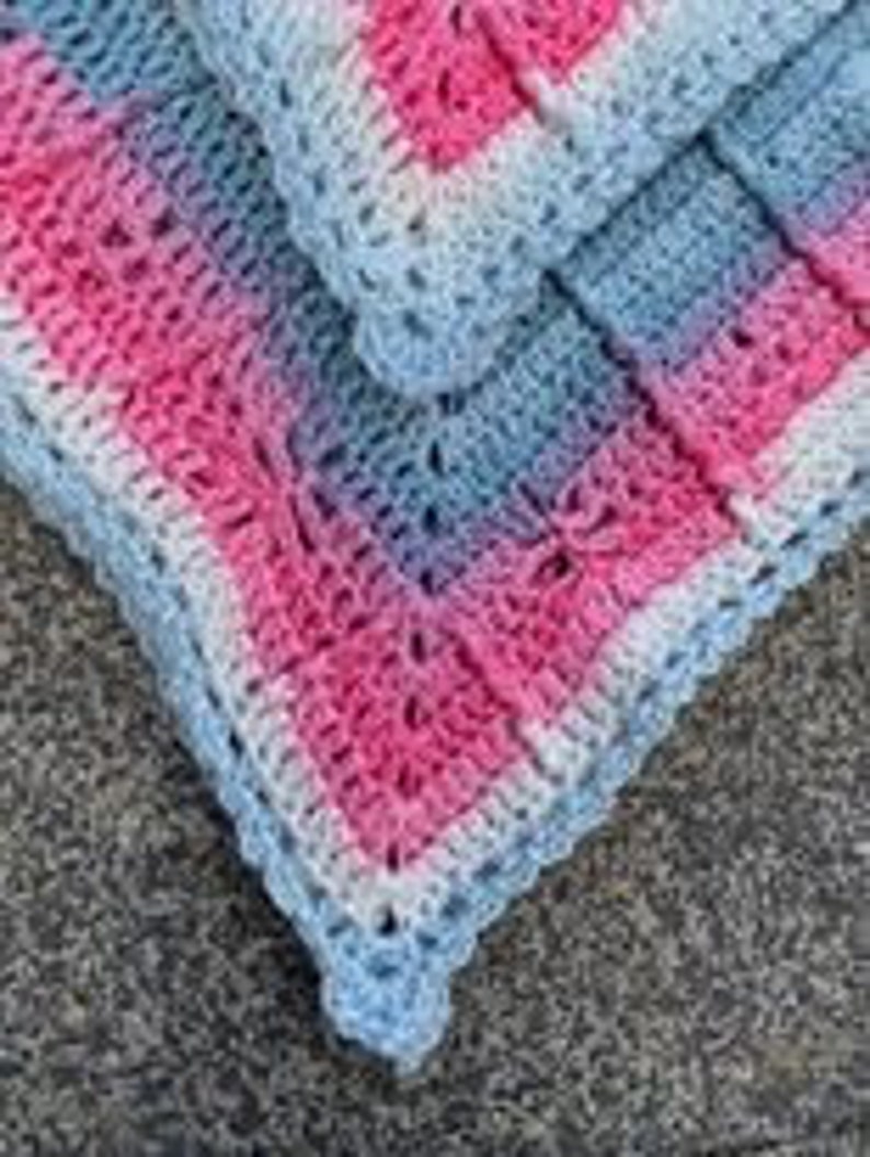 May include: A crocheted baby blanket with a pink, white and blue ombre pattern. The blanket has a delicate crocheted border.