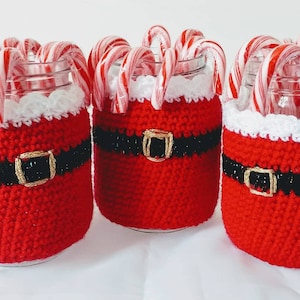 May include: Three glass jars decorated with red crochet Santa Claus suits, black belts with gold buckles, and white trim. Each jar is filled with red and white striped candy canes, perfect for holiday decor.