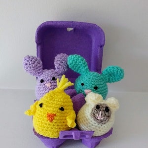 May include: Four crocheted stuffed animals in the shape of a bunny, chick, lamb, and another bunny. The animals are in a purple egg carton.