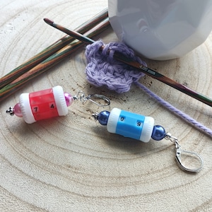 Puede incluir: Dos contadores de puntos coloridos con números negros, uno rojo y otro azul, con ganchos de pendientes plateados y cuentas azul oscuro. Los contadores están sobre una superficie de madera.