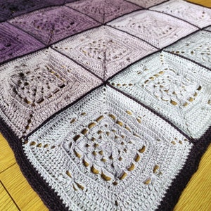 May include: A crocheted blanket with a gradient of purple and white squares. The blanket is made with a delicate stitch pattern and has a dark purple border.
