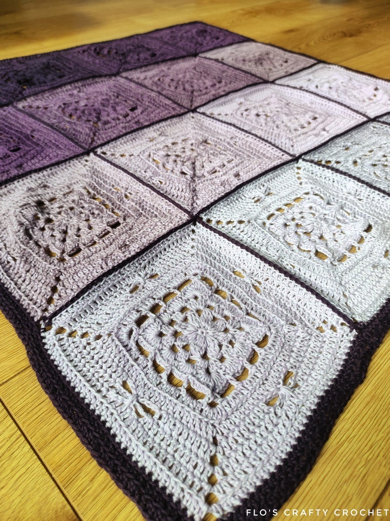 May include: A crocheted blanket with a gradient of purple and white squares. The blanket is made with a delicate stitch pattern and has a dark purple border.