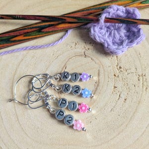 May include: Close-up of crochet stitch markers with silver metal clasps, each with lettered beads and small flower charms in pink, blue, and purple. Knitting needles and a lavender crocheted flower are also visible. The text "FLO'S CRAFTY CROCHET" is at the bottom.