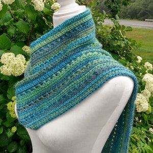 May include: A crocheted scarf in shades of blue and green, with a lacy pattern. The scarf is draped over a white mannequin, and the background is a blurry image of green foliage.