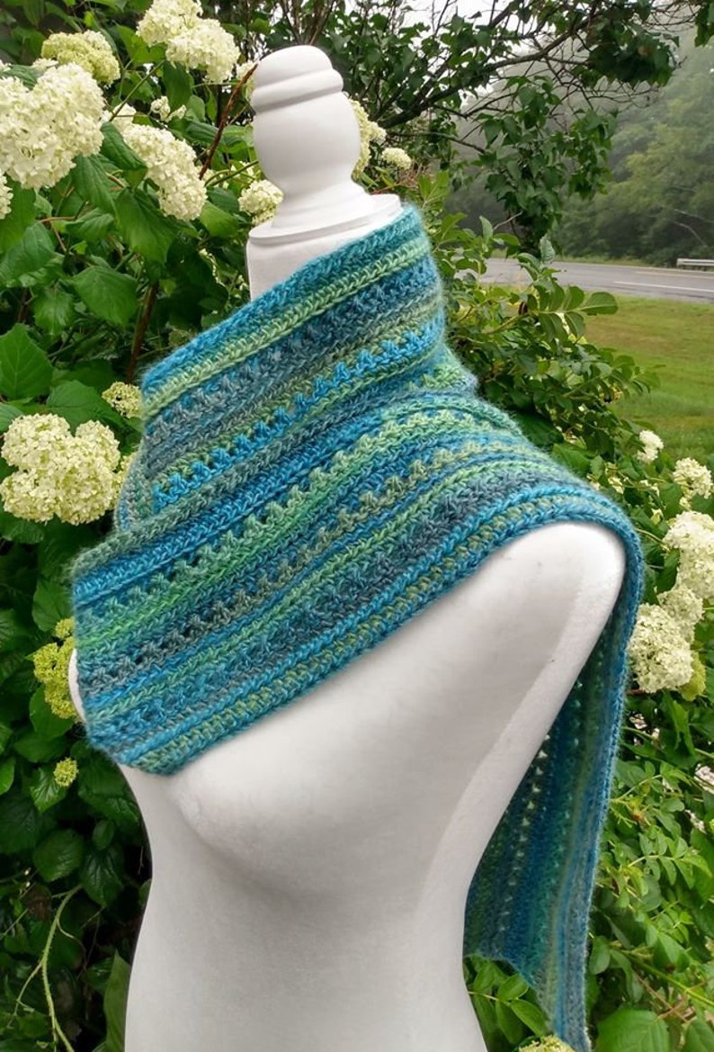 May include: A crocheted scarf in shades of blue and green, with a lacy pattern. The scarf is draped over a white mannequin, and the background is a blurry image of green foliage.