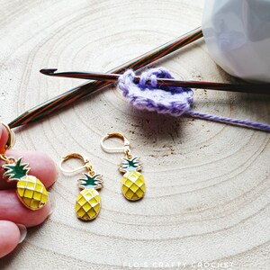 Puede incluir: Dos pendientes de piña dorados con tapas verdes y cuerpos amarillos. Los pendientes están sobre una superficie de madera con una pieza de ganchillo morada y un gancho de ganchillo de madera.