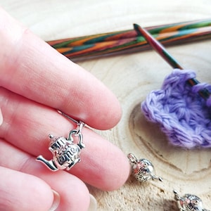 Könnte beinhalten: Silberner Teekannen-Maschenmarkierer mit detailliertem Design. Der Markierer wird in einer Hand gehalten, im Hintergrund ein lilafarbenes Häkelprojekt und Stricknadeln.