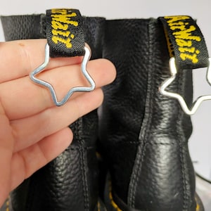 Puede incluir: Botas de cuero negro con costuras amarillas y un lazo de metal en forma de estrella en cada bota. El lazo está unido a una correa negra con texto amarillo que dice "Dr. Martens".