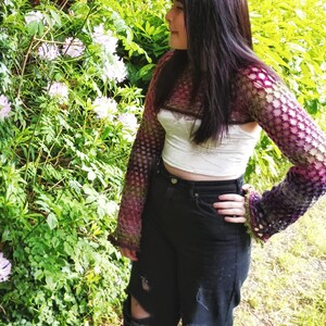 Peut inclure: Une personne portant un haut en crochet violet et vert &agrave; manches longues et un haut court blanc en dessous. La personne porte un jean noir avec des d&eacute;chirures aux genoux et se tient devant un buisson avec des fleurs roses.