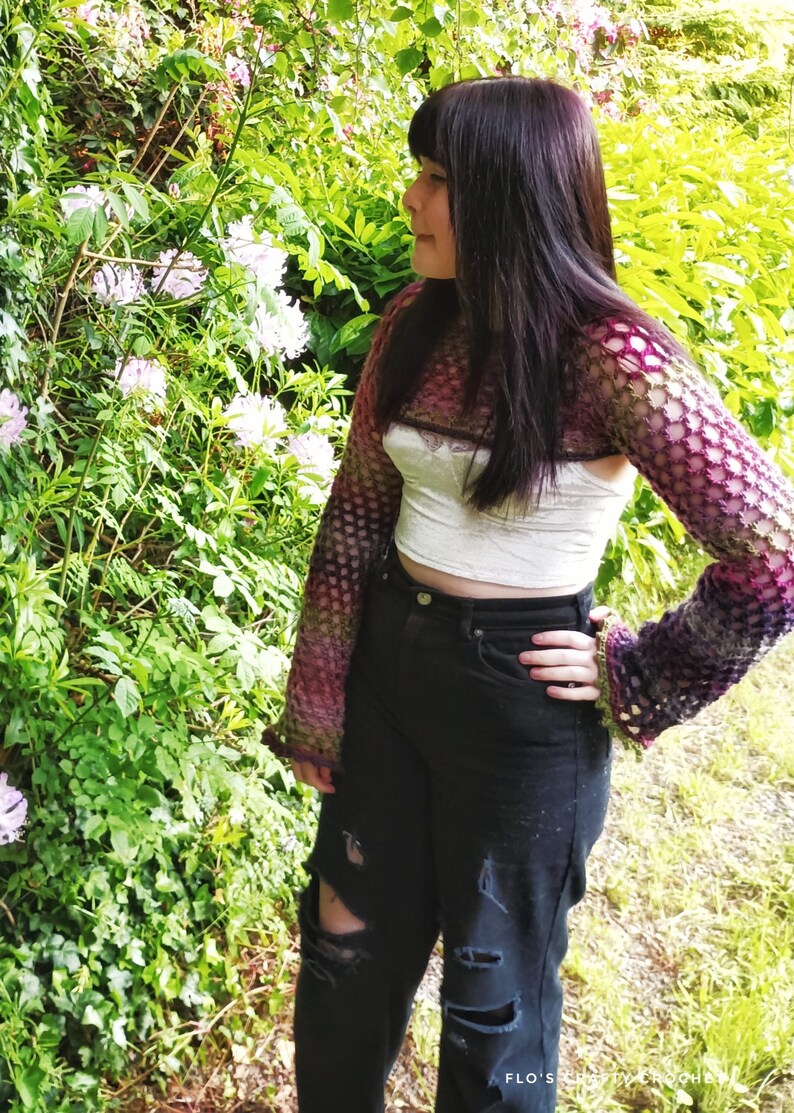 Peut inclure: Une personne portant un haut en crochet violet et vert &agrave; manches longues et un haut court blanc en dessous. La personne porte un jean noir avec des d&eacute;chirures aux genoux et se tient devant un buisson avec des fleurs roses.