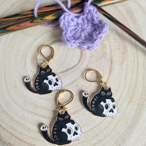 May include: Three black and white cat-shaped earrings with skull designs, gold-coloured ear wires, and a crochet project in progress. The earrings are on a wooden surface, with crochet hooks and a lilac crocheted piece.