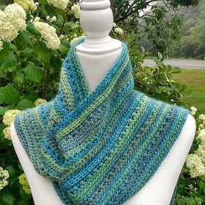 May include: A crocheted scarf in shades of blue, green, and gray. The scarf is displayed on a white mannequin against a backdrop of green foliage.