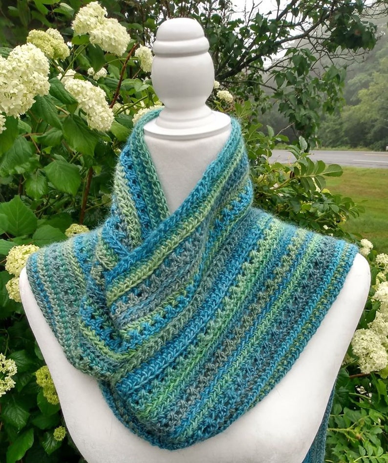 May include: A crocheted scarf in shades of blue, green, and gray. The scarf is displayed on a white mannequin against a backdrop of green foliage.