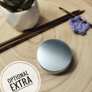 Pu&ograve; includere: Una scatola di metallo argentata con le parole "OPTIONAL EXTRA" stampate su un cerchio bianco. La scatola &egrave; appoggiata su una superficie di legno con un paio di ferri da maglia e un quadrato all'uncinetto viola.