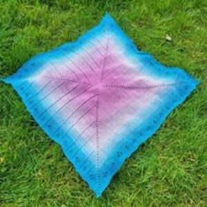 May include: A square crocheted shawl with a gradient of blue, pink and white. The shawl is laid out on a green grassy surface.