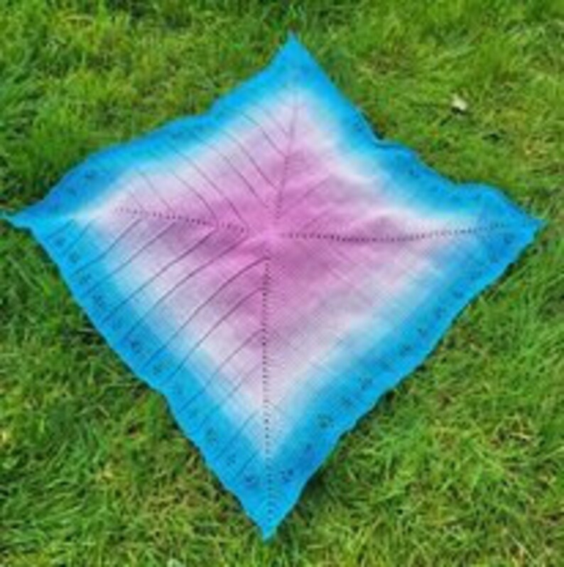May include: A square crocheted shawl with a gradient of blue, pink and white. The shawl is laid out on a green grassy surface.