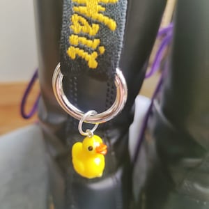 Op de afbeelding: Zwarte leren laarzen met een gele rubberen eendjescharme aan een zilveren ring. De laarzen hebben een zwarte band met gele stiksels waarop "Dr. Martens" staat.
