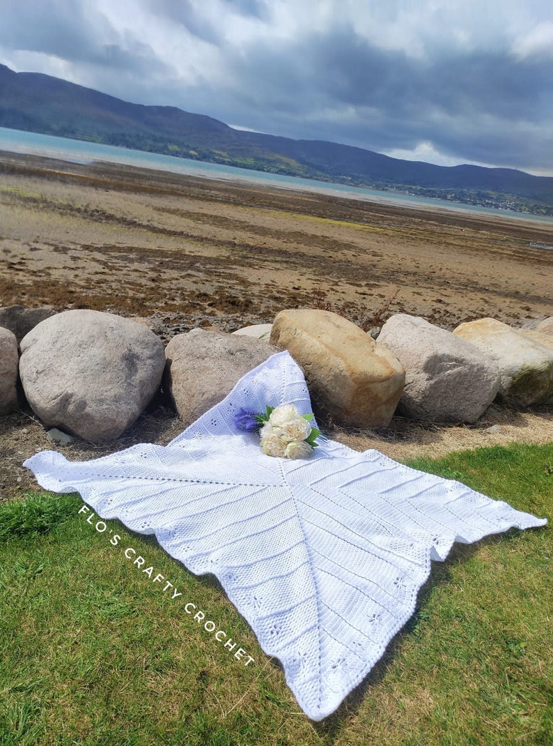 May include: White crochet baby blanket with a scalloped edge, laid out on green grass with a bouquet of white flowers. The blanket has the text "Flo's Crafty Crochet" written on it.