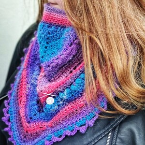 Peut inclure: Un col crochet&eacute; color&eacute; dans les tons de violet, rose et bleu. L'&eacute;charpe faite &agrave; la main pr&eacute;sente un bord festonn&eacute; et une fermeture boutonn&eacute;e. Le col est port&eacute; sur une veste noire.