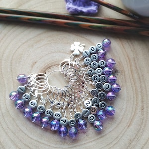 May include: A set of 10 silver stitch markers with purple iridescent beads. Each marker has a number from 1 to 10 printed on it. The markers are arranged in a circle with the beads facing outwards. The markers are on a wooden surface.