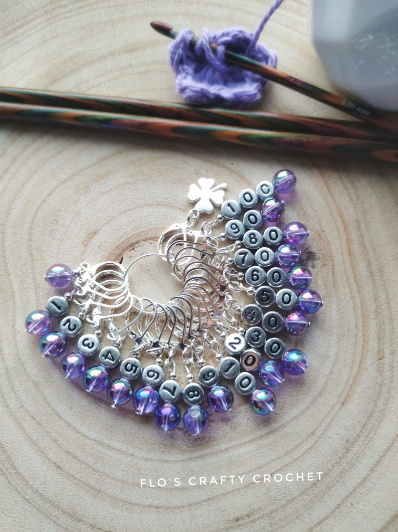 May include: A set of 10 silver stitch markers with purple iridescent beads. Each marker has a number from 1 to 10 printed on it. The markers are arranged in a circle with the beads facing outwards. The markers are on a wooden surface.