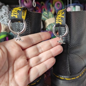 May include: A pair of black leather boots featuring yellow stitching along the sole and a black and yellow heel pull tab with the text AirWair. Each boot has a silver metal ring attached to the pull tab, each holding a small dinosaur charm.