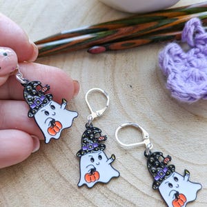 Op de afbeelding: Drie Halloween-geestoorbellen met zilveren haken. Elke witte geest draagt een zwarte en paarse heksenhoed en houdt een oranje pompoen vast. De oorbellen worden gepresenteerd op een houten oppervlak.