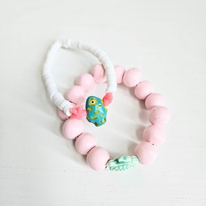 Könnte beinhalten: Zwei Armbänder, eines weiß und eines rosa. Das rosa Armband hat einen blauen und gelben Fisch-Anhänger und einen grünen Schildkröten-Anhänger.