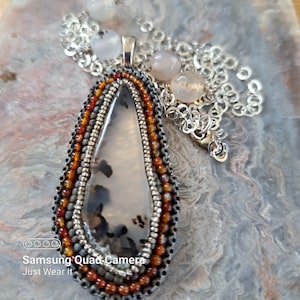 May include: A handcrafted pendant necklace with a teardrop-shaped stone displaying black and grey patterns. The stone is framed with layers of orange, grey, and black beads. The necklace has a silver chain and stone accents. The text "Just Wear It" is visible.