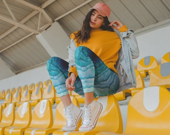 Puede incluir: Una mujer con una sudadera amarilla, una chaqueta vaquera y una gorra de béisbol rosa se sienta en las gradas de un estadio amarillo. Lleva leggings con estampado azul y blanco y zapatillas blancas.