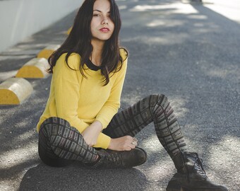 May include: A woman wearing a yellow jumper and black and white patterned leggings sits on a grey asphalt surface. She is wearing black combat boots.