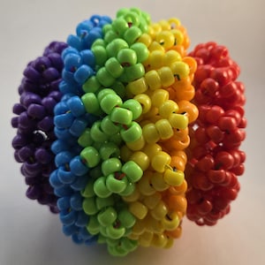 May include: A colorful, spherical object composed of tightly wound, rainbow-colored beads. The beads are arranged in bands of red, orange, yellow, green, blue, and purple. The beads have a spiral shape and are connected to form a ball.
