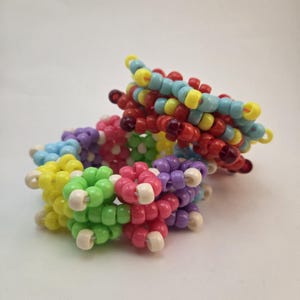 May include: A multi-colored beaded bracelet constructed from small plastic pony beads in shades of yellow, lime green, pink, purple, and light blue, arranged in a repeating floral pattern with white beads at the center of each flower shape.