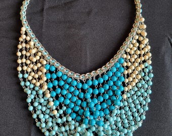May include: A layered necklace with a silver chain and blue and cream colored beads. The beads are arranged in a graduated pattern, with the largest beads at the center of the necklace and the smallest beads at the edges.