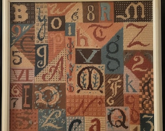 May include: A framed cross-stitch sampler featuring a patchwork design of letters and numbers in various fonts and colors, including brown, blue, and orange. The artwork is displayed in a light-colored wooden frame.