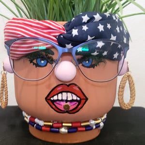 May include: A terracotta flower pot painted with a face design, featuring blue-framed glasses, a pink nose, and red lips. The pot is adorned with a red and white striped headband, a blue star-patterned scarf, and beaded necklaces. The pot has faux eyelashes and earrings.