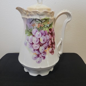 May include: White ceramic pitcher with a lid, featuring a hand-painted design of purple grapes and green leaves. The pitcher has a curved handle and a spout, with a scalloped rim and a cream-colored lid.