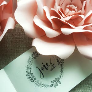 May include: A white card with black calligraphy that reads "joie de vivre" inside a wreath of leaves. The card is lying on top of a large, pink paper flower.