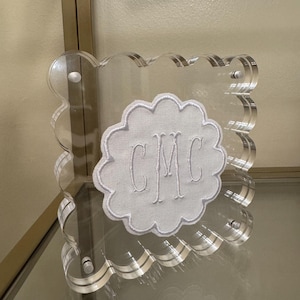 May include: Clear acrylic frame with a scalloped edge. A white fabric applique with the embroidered initials "CMC" is centered within the frame. The frame is displayed on a glass shelf.