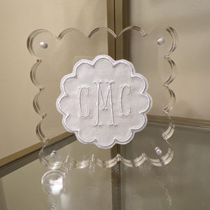 May include: Clear acrylic monogrammed plaque with a scalloped edge. The white embroidered monogram "CMC" is centered on a white fabric cloud shape. The plaque is secured with four silver-colored fasteners.