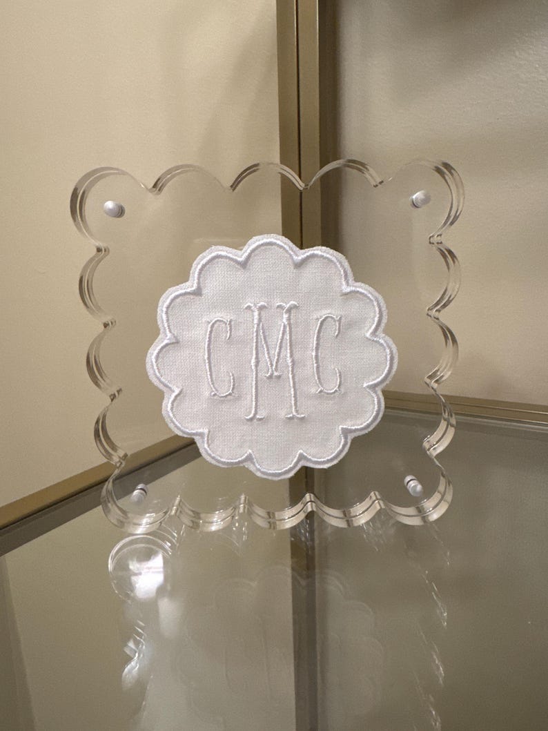 May include: Clear acrylic monogrammed plaque with a scalloped edge. The white embroidered monogram "CMC" is centered on a white fabric cloud shape. The plaque is secured with four silver-colored fasteners.