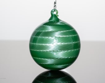 May include: A green glass ornament with a swirling, textured pattern. The ornament is round with a loop at the top for hanging. The glass has a glossy finish and reflects light.