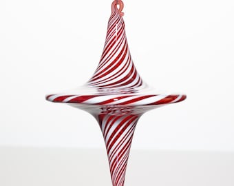May include: A hand-blown glass ornament featuring a spinning top shape with a pointed top and bottom. The surface is decorated with a continuous pattern of swirling red and white diagonal stripes that wrap around the entire glass body.