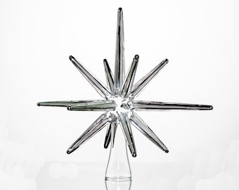 May include: A silver glass star tree topper with a pointed design. The star has multiple points and is made of clear glass with a reflective finish.