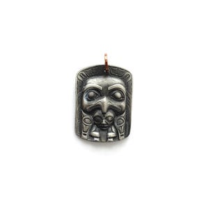 Peut inclure: Pendentif rectangulaire argenté avec un motif de totem amérindien détaillé. Le pendentif présente un visage central avec des traits stylisés, entouré de gravures complexes. Une boucle de couleur cuivre est fixée en haut pour la suspension.