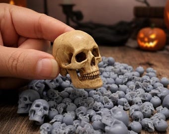 May include: A tan-colored miniature skull held above a pile of small, gray skulls. The detailed skull has visible teeth and eye sockets. The background includes lit candles and carved pumpkins, suggesting a Halloween theme.