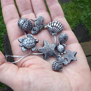 Puede incluir: Una colección de dijes plateados con temática marina, que incluye estrellas de mar, caballitos de mar, tortugas y conchas. Cada dije está intrincadamente detallado y unido a un fino cordón plateado. Los dijes se muestran en una mano abierta.